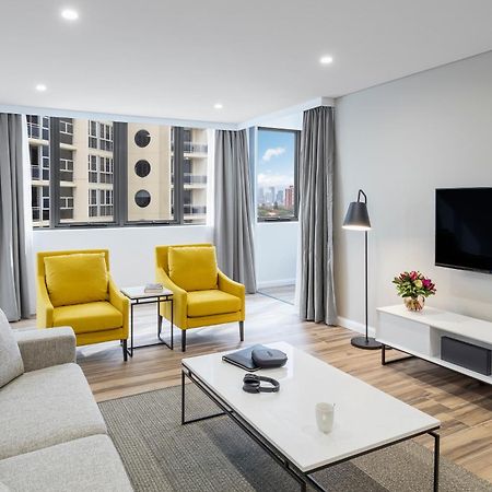 Meriton Suites Bondi Junction Sydney Exterior foto The photo shows a modern, well-lit living room. It features a light gray sofa with a textured fabric. There are two bright yellow armchairs positioned near a window, which allows natural light to enter the space. The room has light gray curtains and 