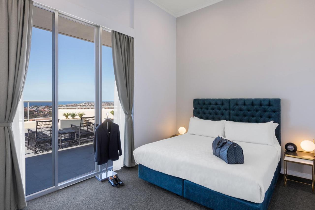 Meriton Suites Bondi Junction Sydney Exterior foto The photo shows a stylish bedroom featuring a large bed with a tufted blue headboard. There's a neatly organized outfit consisting of a suit hanging on a stand, along with shoes placed on the floor. The room has large windows that provide a view of t