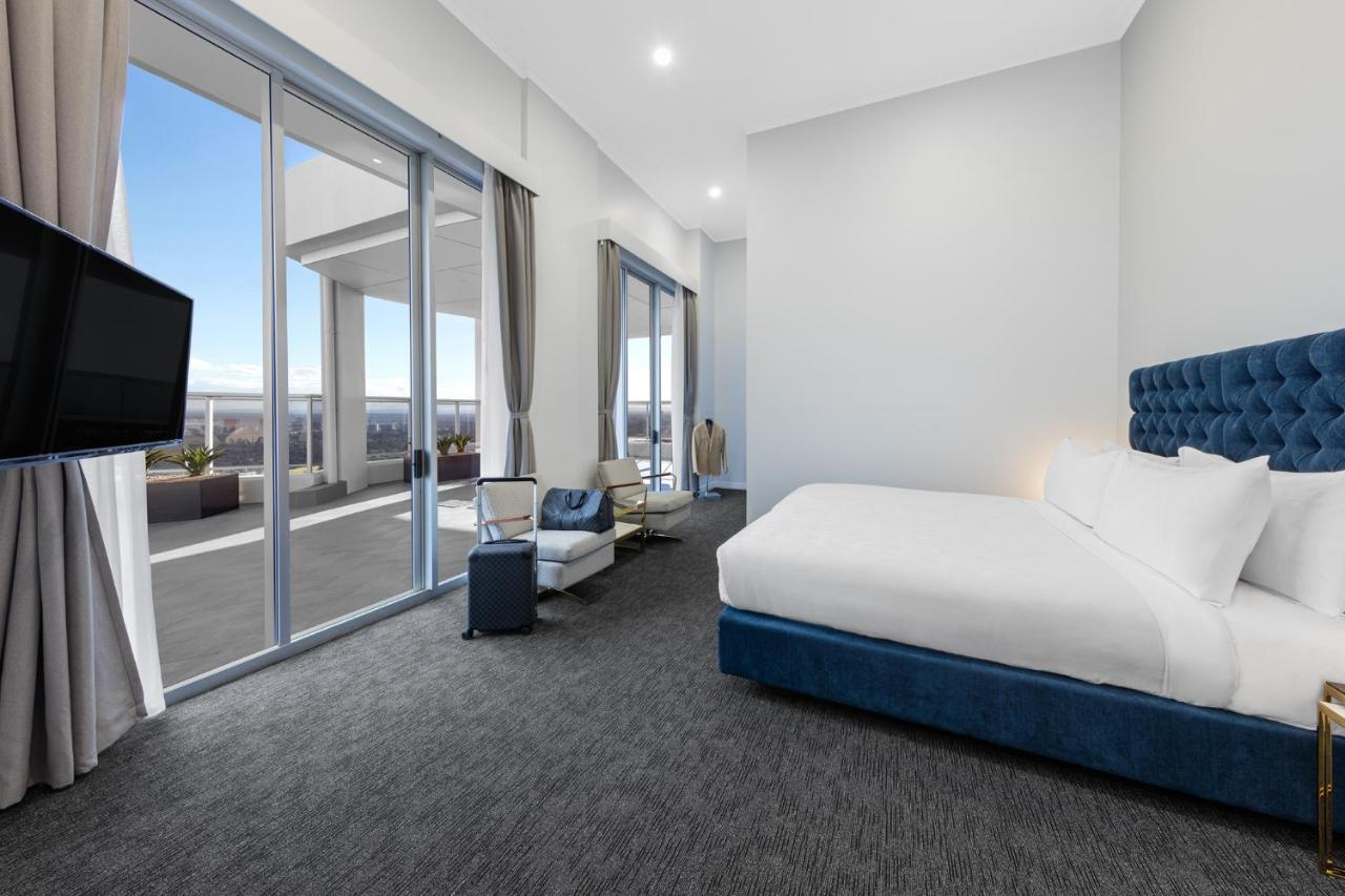Meriton Suites Bondi Junction Sydney Exterior foto The photo shows a modern and spacious hotel room. It features a large bed with a blue headboard, a flat-screen TV mounted on the wall, and a cozy seating area with a chair and small table. The room has large windows that offer natural light and a vie