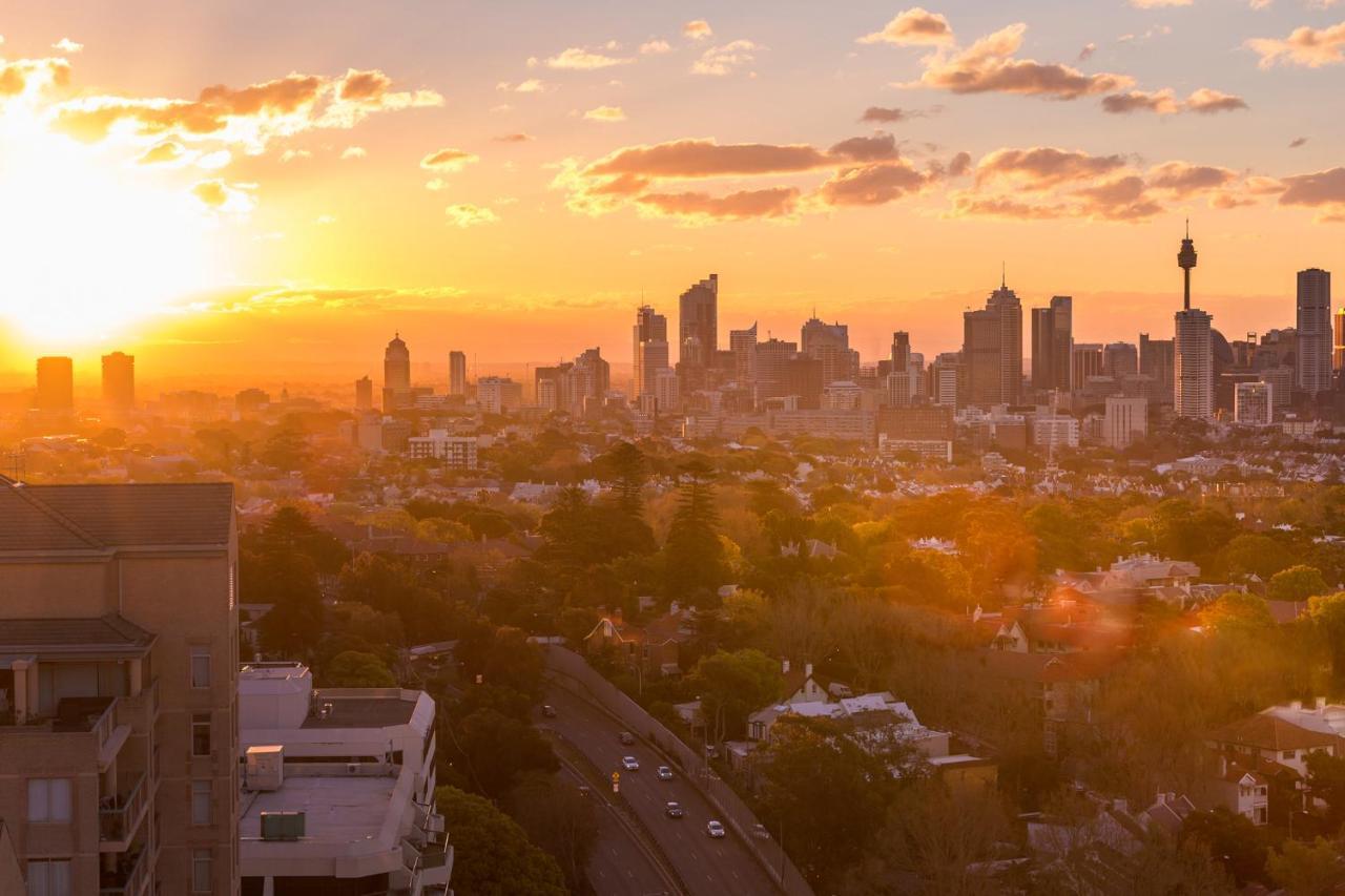 Meriton Suites Bondi Junction Sydney Exterior foto The photo shows a panoramic view of a city skyline at sunset. The sun is low on the horizon, casting a warm golden light across the buildings. Tall skyscrapers are silhouetted against the colorful sky, which features hues of orange, pink, and yellow,