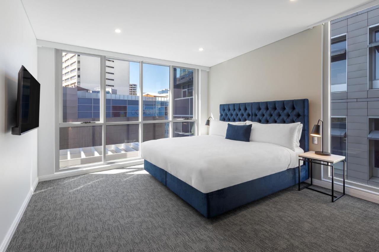 Meriton Suites Bondi Junction Sydney Exterior foto The image shows a modern hotel room with a large, comfortable bed featuring a blue upholstered headboard. The room has large windows that allow natural light to enter, providing a good view of the surrounding buildings. There is a small bedside table
