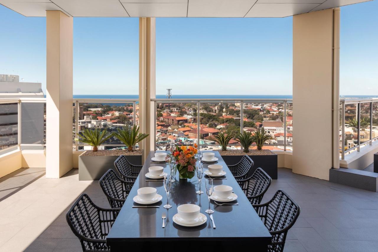 Meriton Suites Bondi Junction Sydney Exterior foto The photo shows a modern outdoor dining area situated on a balcony or terrace with a stunning view. There is a long rectangular table set with plates, glasses, and cutlery, ready for a meal. The table is surrounded by sleek, black wicker chairs. In t