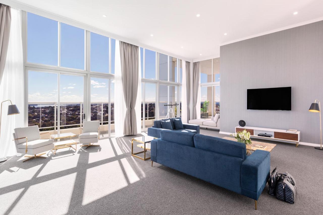 Meriton Suites Bondi Junction Sydney Exterior foto The photo shows a bright and modern living room with large floor-to-ceiling windows that provide a view of the outdoors. The space features a blue sofa in the center, complemented by light-colored armchairs nearby. There are light curtains hanging by
