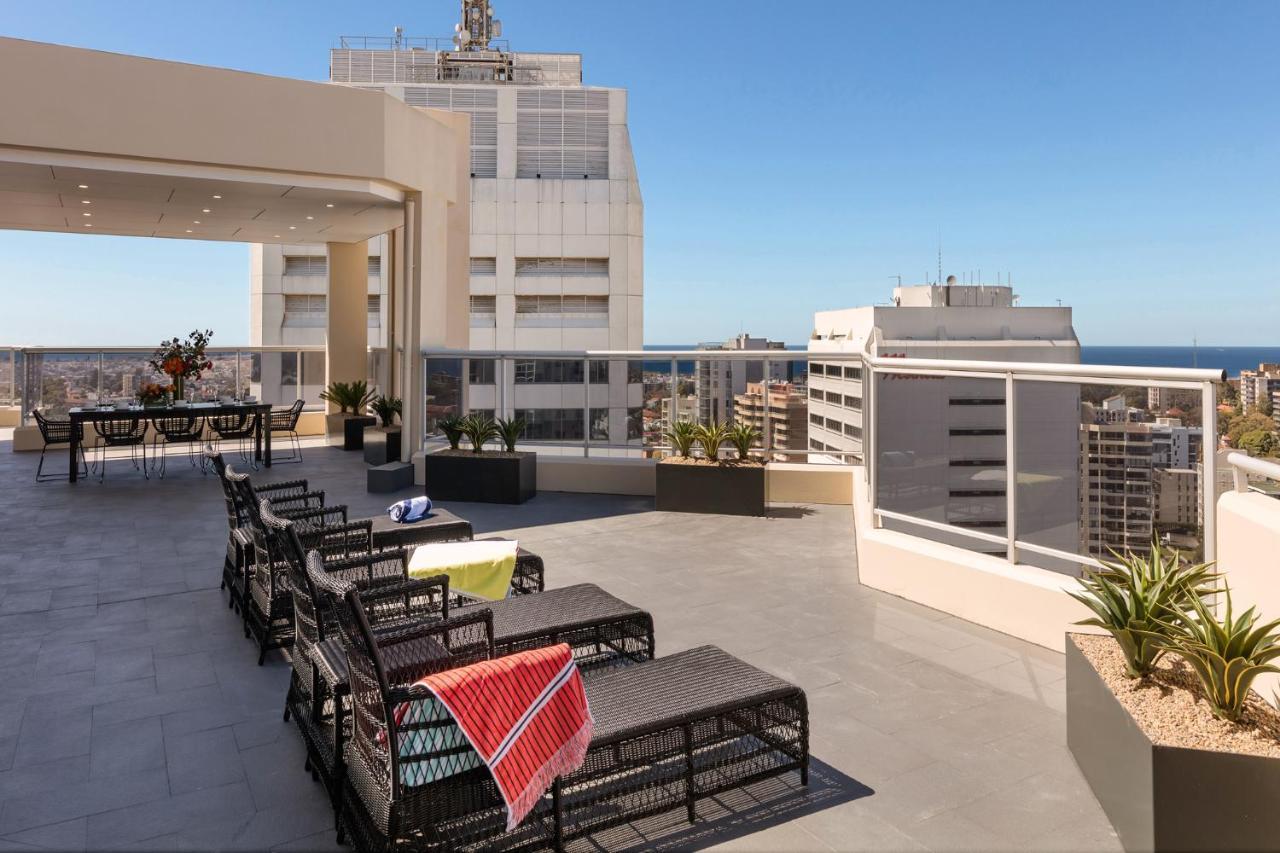 Meriton Suites Bondi Junction Sydney Exterior foto The photo shows a rooftop terrace with a modern design. There are several outdoor seating arrangements featuring black wicker lounge chairs. Some of the chairs have colorful blankets draped over them. The terrace is surrounded by a railing and has po