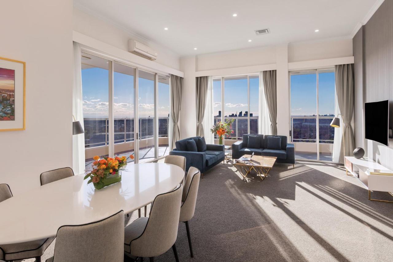 Meriton Suites Bondi Junction Sydney Exterior foto The photo shows a modern and stylish living space with large windows allowing natural light to fill the room. In the foreground, there is a round dining table with several chairs around it, adorned with a floral centerpiece. The seating area features