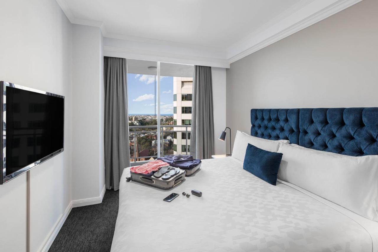 Meriton Suites Bondi Junction Sydney Exterior foto The photo shows a modern hotel room featuring a neatly made bed with a blue tufted headboard and a white duvet. There's a small decorative pillow on the bed, and a piece of luggage is placed at the foot with clothes partially unpacked. A television i