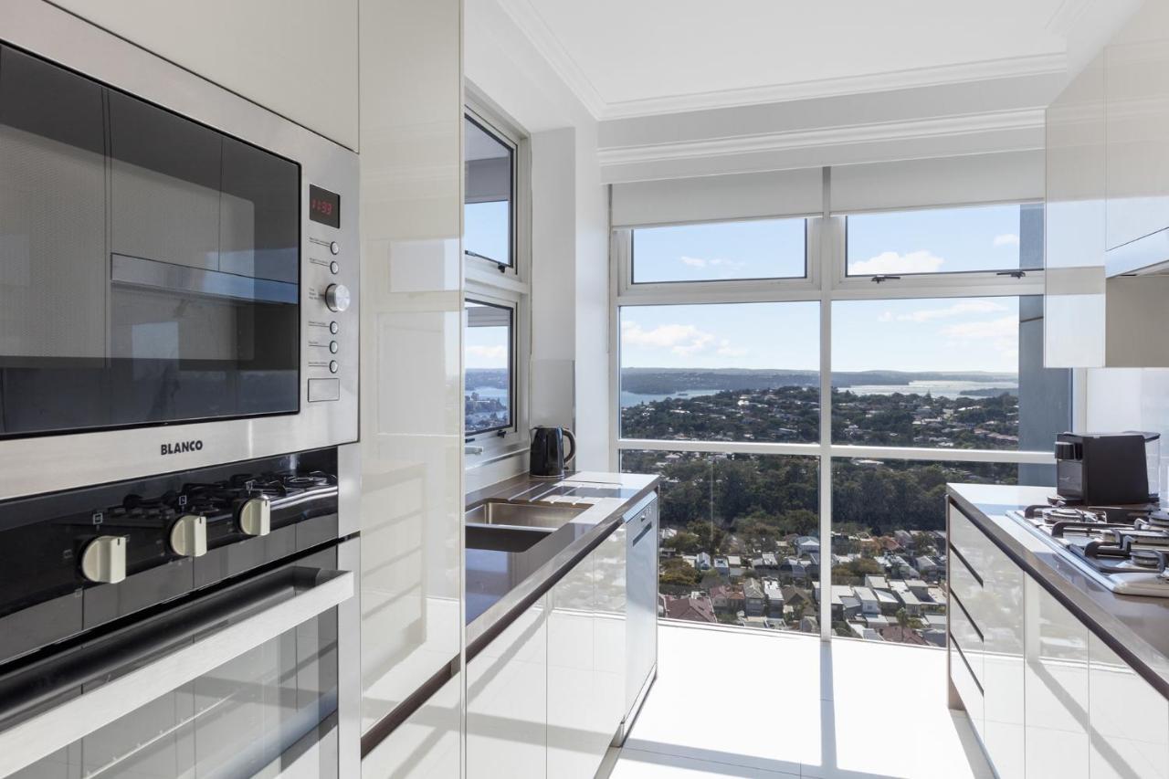 Meriton Suites Bondi Junction Sydney Exterior foto The photo shows a modern kitchen with sleek, shiny white cabinets and a large window that offers a panoramic view of the outside landscape. The kitchen features built-in appliances, including an oven and a stove, with a stylish countertop. The spacio