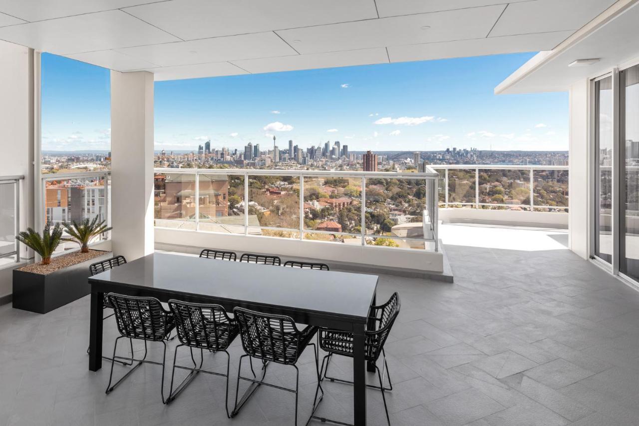 Meriton Suites Bondi Junction Sydney Exterior foto The photo shows a modern outdoor balcony or terrace space with a dining table and chairs. The table is black and has a minimalist design, surrounded by stylish chairs. In the background, there is a stunning view of a city skyline under a clear blue s