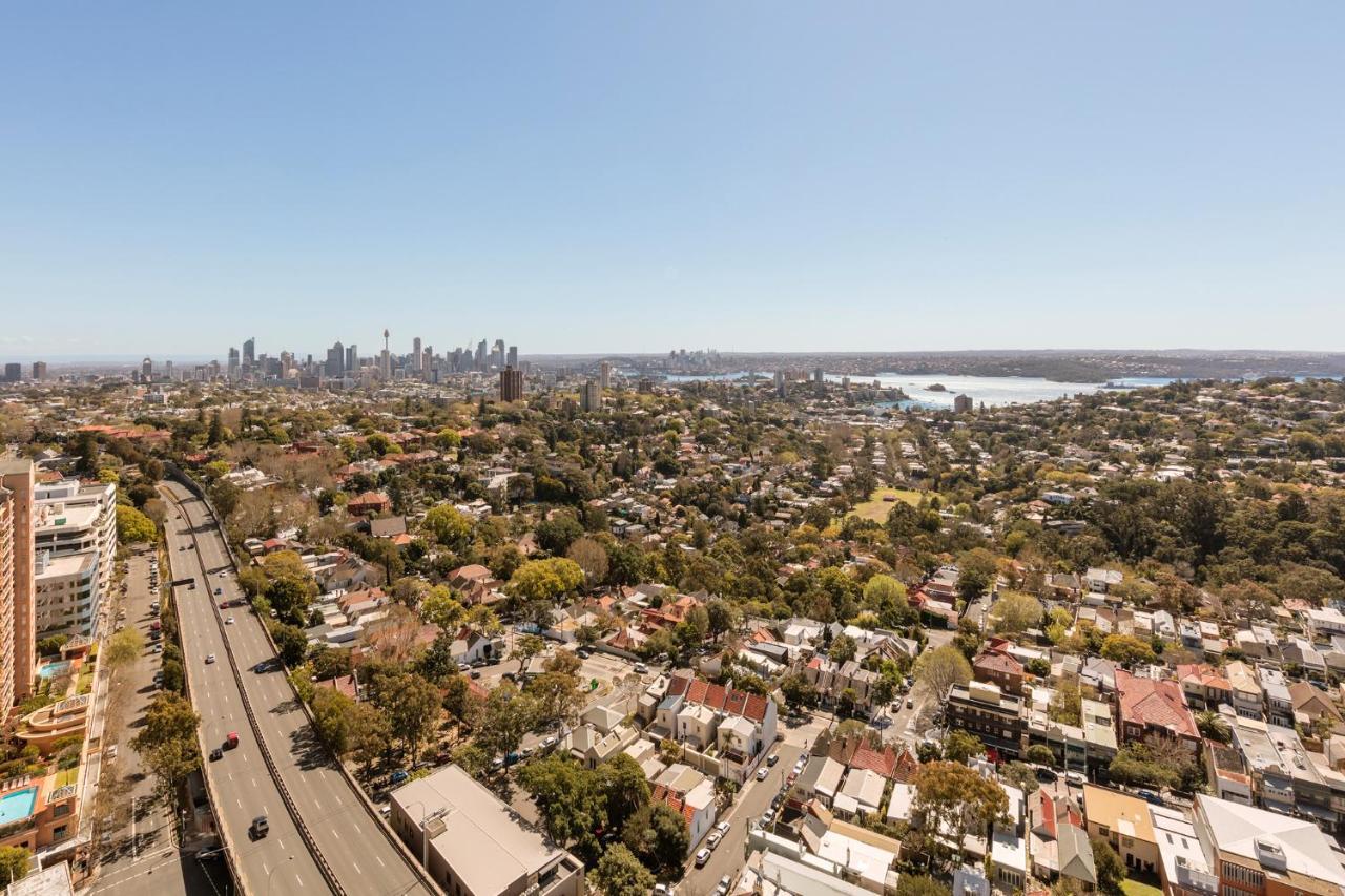 Meriton Suites Bondi Junction Sydney Exterior foto The photo captures a panoramic view of a vibrant urban landscape. In the foreground, there are residential areas with various houses and greenery, showcasing a suburban environment. In the background, the skyline of a city can be seen, featuring tall