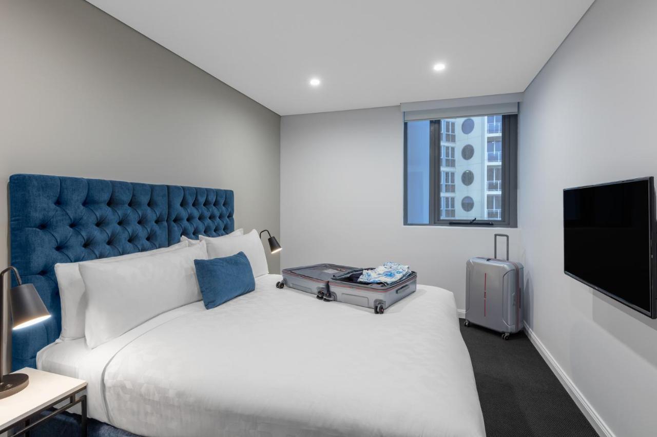 Meriton Suites Bondi Junction Sydney Exterior foto The photo shows a modern hotel room featuring a neatly made bed with a blue tufted headboard. There is a small blue decorative pillow on the bed, and the bedding is white. Next to the bed, there is a nightstand with a lamp. A suitcase is partially op