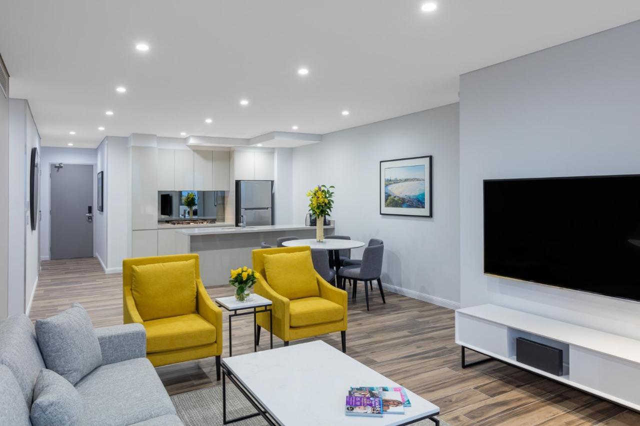 Meriton Suites Bondi Junction Sydney Exterior foto The photo shows a modern living space that includes a cozy sitting area and a dining area. The sitting area features two bright yellow chairs and a light gray sofa, arranged around a coffee table. There are decorative items on the table, including fl