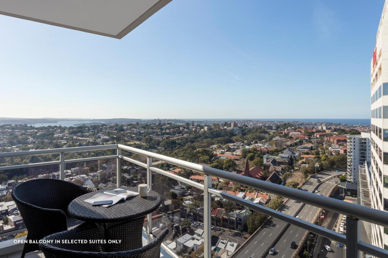 Meriton Suites Bondi Junction Sydney Exterior foto The photo shows a balcony with a view over a cityscape that includes buildings and greenery, extending towards the horizon where the sea is visible. The balcony features a small round table and two black chairs, suggesting a space for relaxation or d