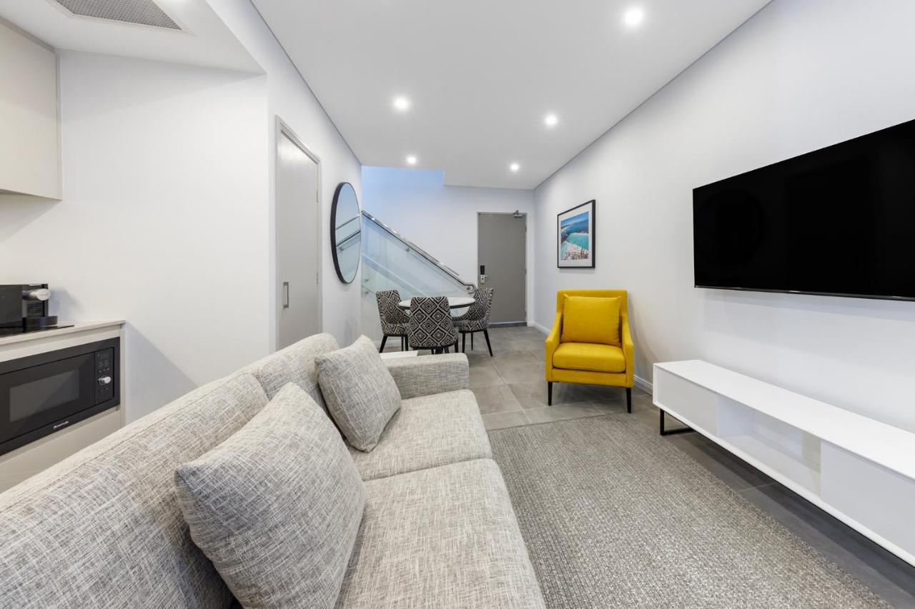 Meriton Suites Bondi Junction Sydney Exterior foto The photo shows a modern and stylish living space. It features a light-colored sofa with cushions, positioned to face a wall-mounted flat-screen TV. There's a yellow accent chair nearby that adds a pop of color to the room. The décor is minimalistic,
