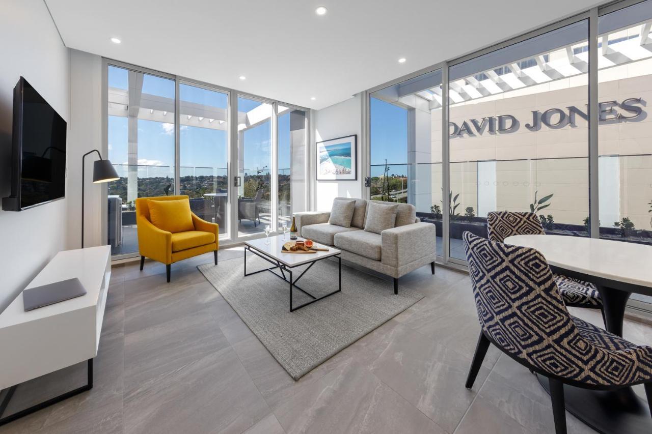 Meriton Suites Bondi Junction Sydney Exterior foto The photo shows a modern living space that features large windows allowing ample natural light. The room is furnished with a gray sofa and a bright yellow armchair, along with a coffee table in between. There’s a decorative bowl with fruits on the ta