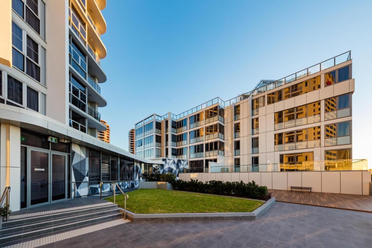 Meriton Suites Bondi Junction Sydney Exterior foto The photo showcases a modern residential area featuring contemporary buildings. In the foreground, there is a landscaped green space surrounded by well-designed architecture. The buildings have large glass windows, reflecting the light, and feature a