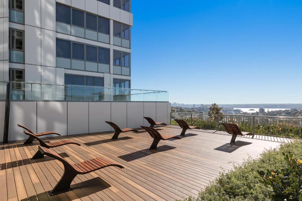 Meriton Suites Bondi Junction Sydney Exterior foto The photo shows a rooftop or outdoor area with a wooden deck. There are several modern lounge chairs arranged neatly, facing a clear sky and a panoramic view of the surrounding landscape. In the background, there is a tall building with large windows