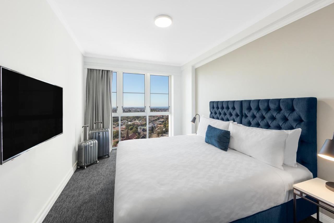 Meriton Suites Bondi Junction Sydney Exterior foto The photo shows a modern hotel room with a large bed featuring a tufted blue headboard. There is a white duvet and a decorative pillow on the bed. A flat-screen television is mounted on the wall opposite the bed. Large windows are covered with gray c