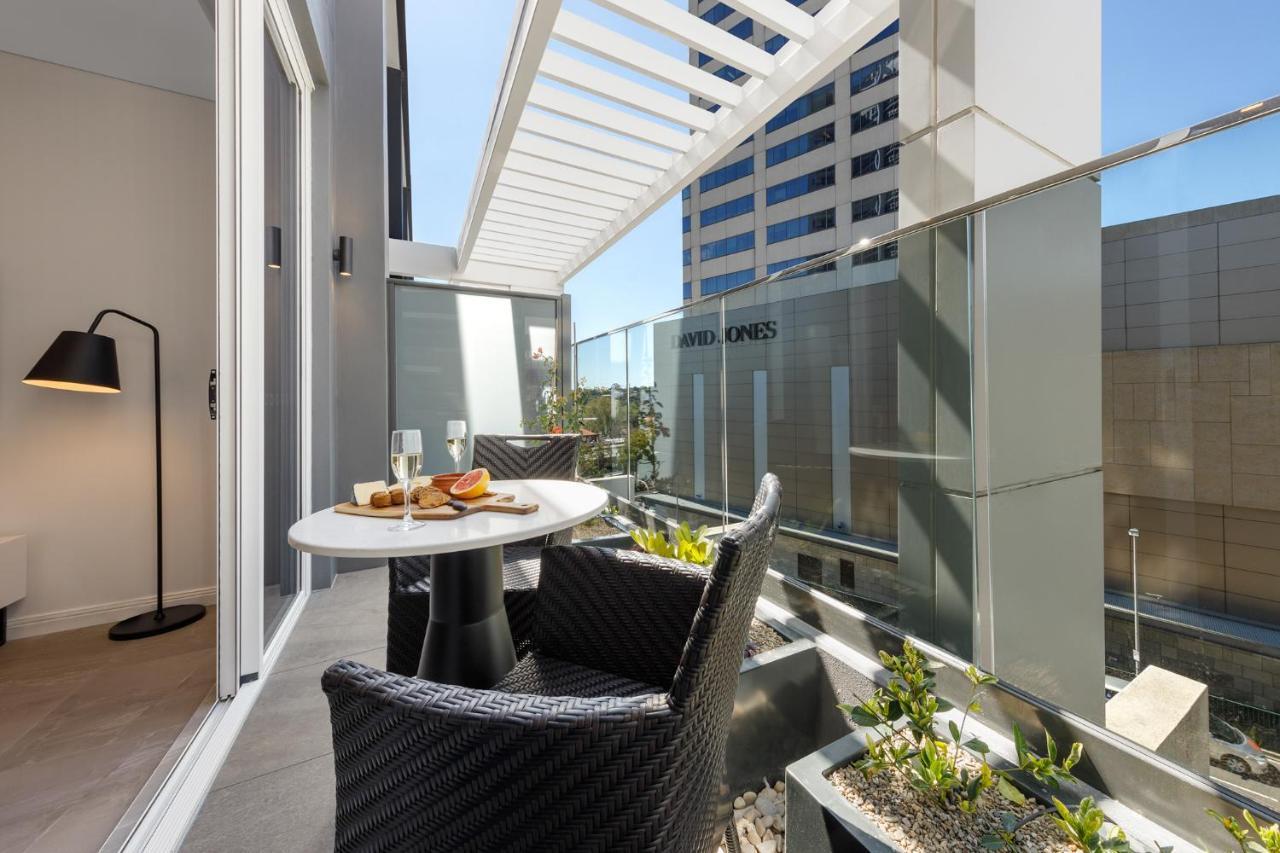Meriton Suites Bondi Junction Sydney Exterior foto The photo shows a modern balcony area with glass railings. It features a small round table set for two, with a plate of food and drinks. There are two stylish wicker chairs positioned around the table. The space is bright and airy, with sunlight stre