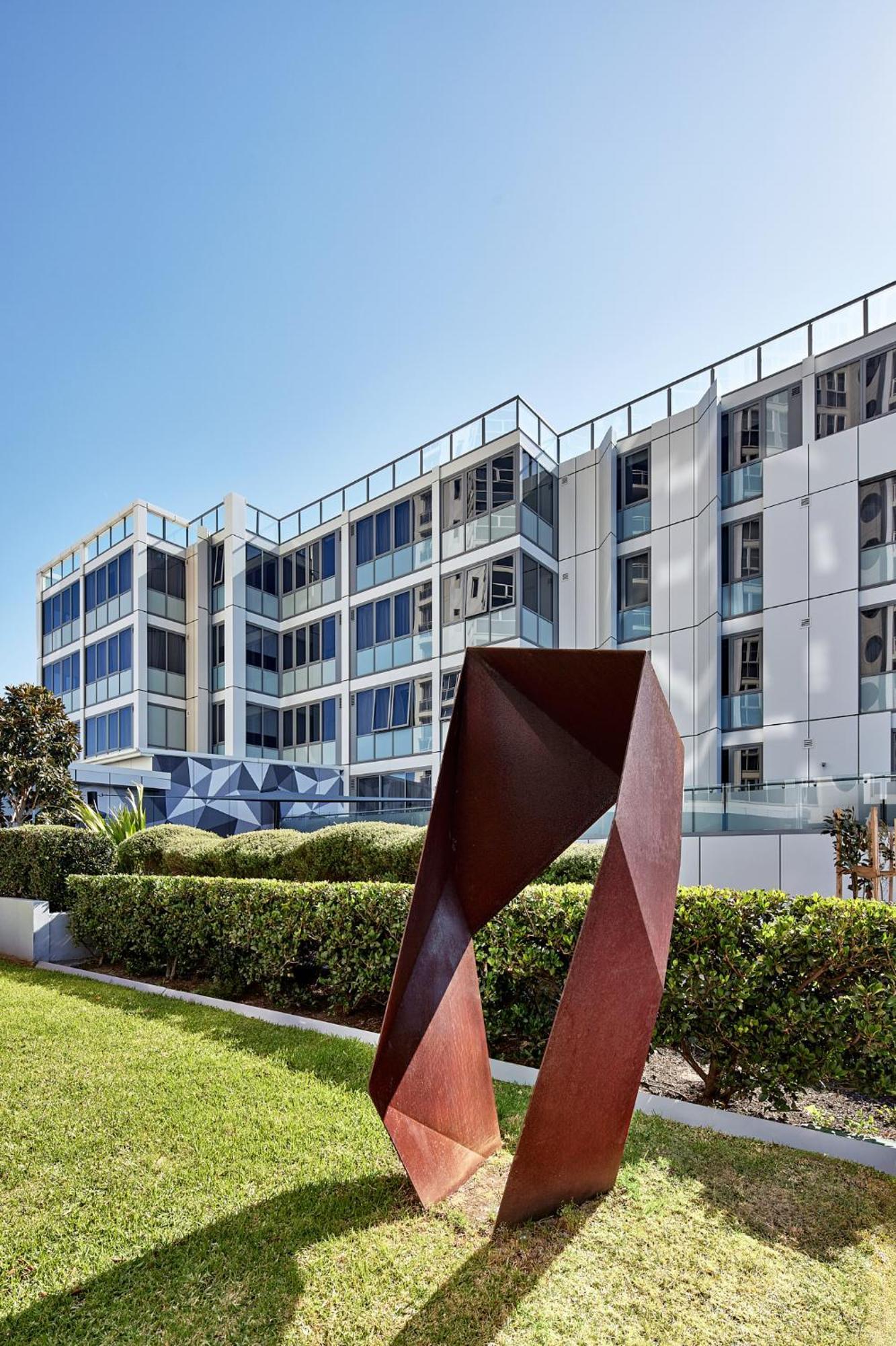 Meriton Suites Bondi Junction Sydney Exterior foto The photo shows a modern building with a sleek white and glass facade, featuring multiple floors and large windows. In the foreground, there is a sculptural piece made of metal, characterized by geometric shapes and a reddish-brown color. The surroun