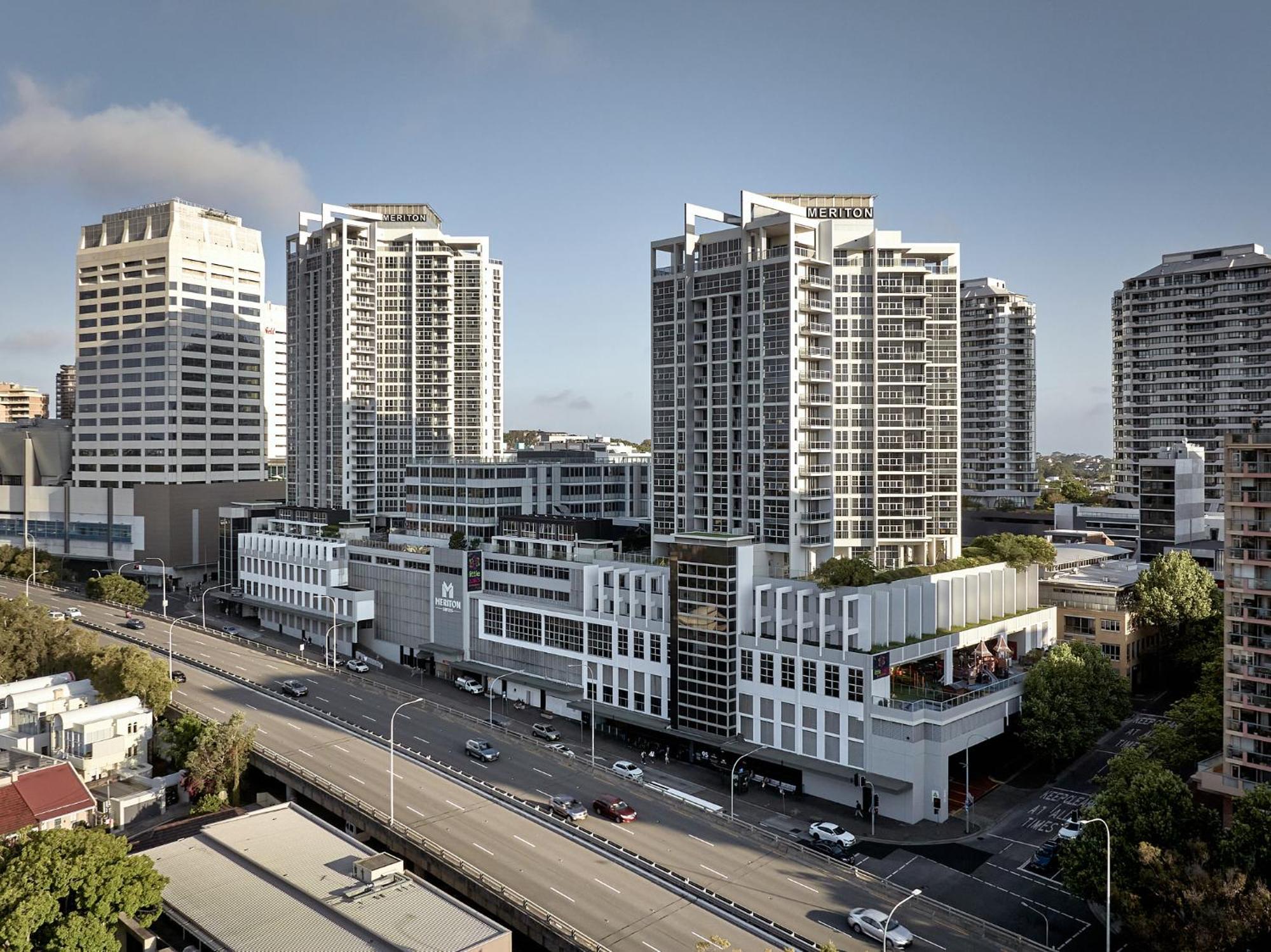 Meriton Suites Bondi Junction Sydney Exterior foto The photo shows a cityscape featuring several tall modern buildings, likely apartments or office complexes. In the foreground, there is a road lined with cars, and the architecture of the buildings varies, with some having sleek glass facades and oth