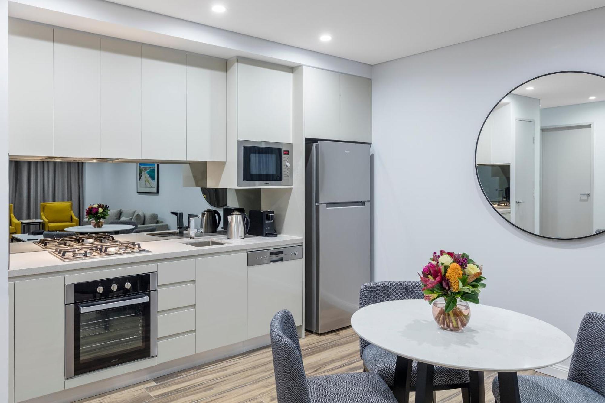 Meriton Suites Bondi Junction Sydney Exterior foto The photo shows a modern kitchen and dining area. The kitchen features light-colored cabinetry with a built-in oven and stovetop, a microwave, and a silver refrigerator. There is a kettle and a coffee maker on the countertop. In the dining area, ther